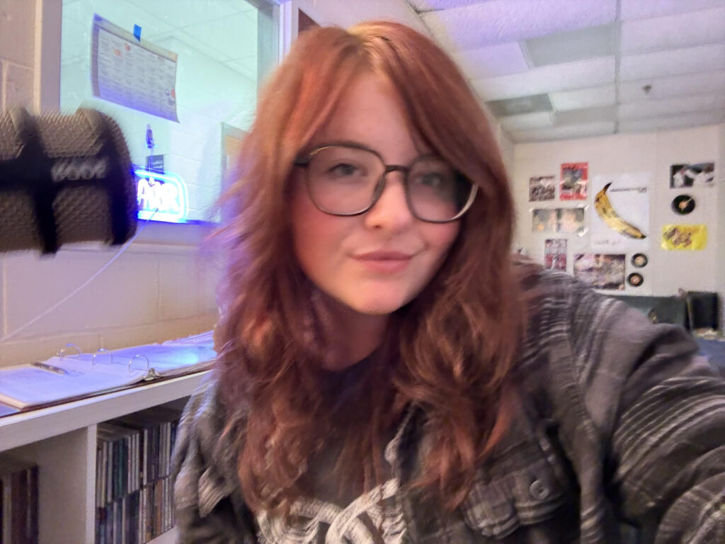 A young woman with shoulder length reddish brown hair and glasses smiling in a college radio station.