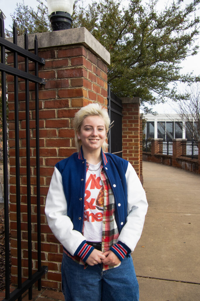 Lee, a young person with a bleach blond mullet, wearing a blue and white letterman jacket, stands with their hands in front of them, grinning at the camera